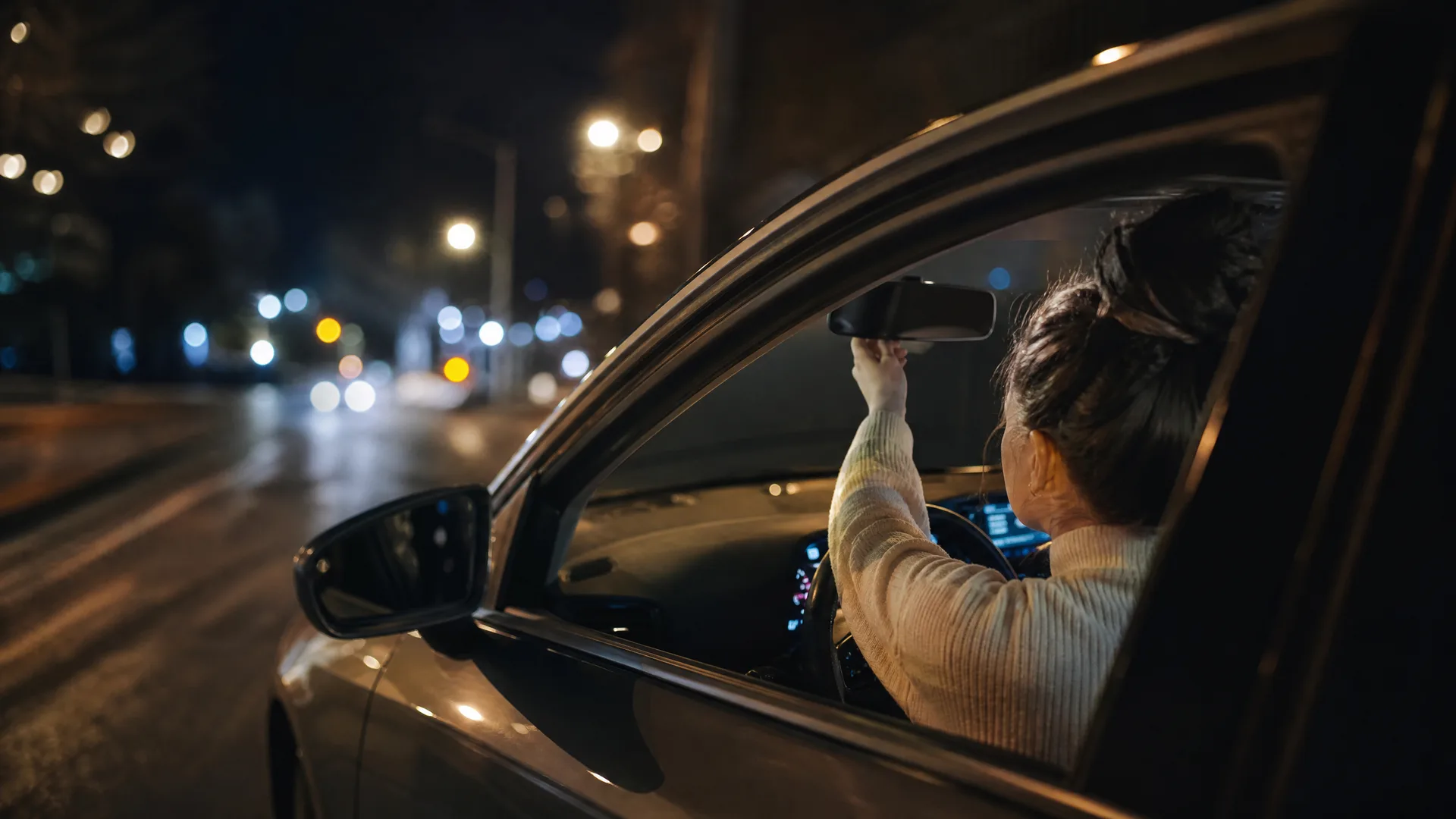 A woman seen from outside the open driver's window of a car at night, reaching up to adjust the rear-view mirror, with blurred street lights in the background.